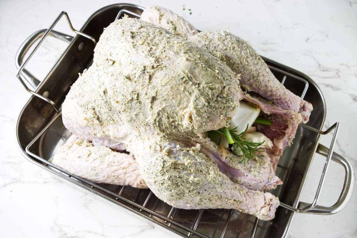 Whole turkey slathered with butter and herbs in a roasting pan.