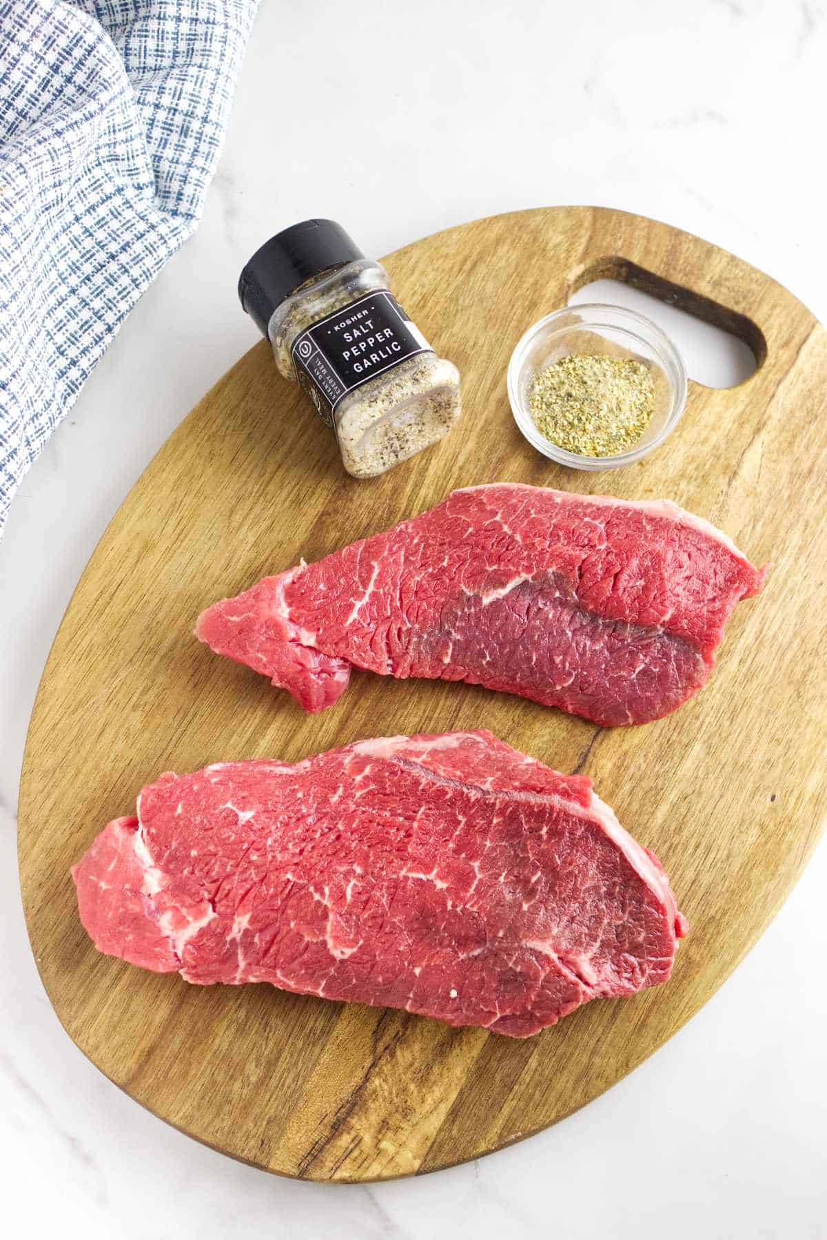 Raw Denver steaks with small bowl of seasonings on a cutting board.