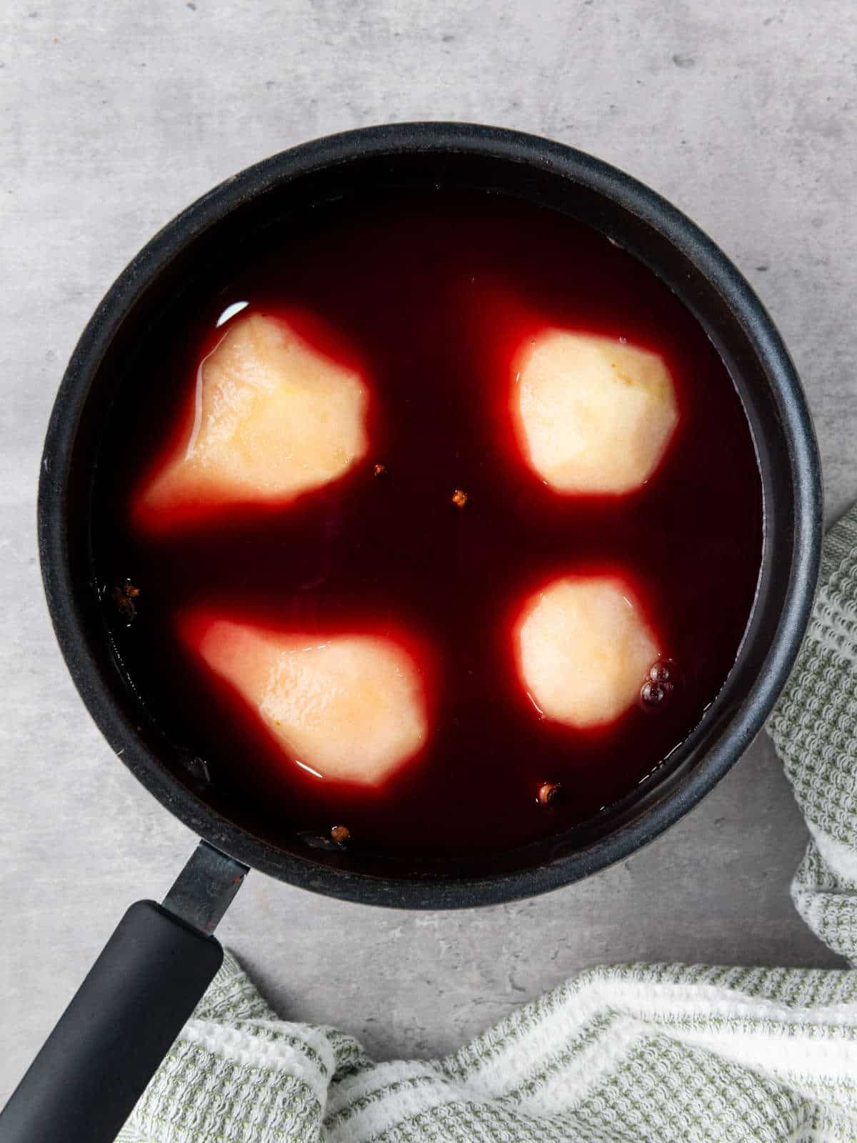 Four peeled pears in a sauce pan with red wine and spices.